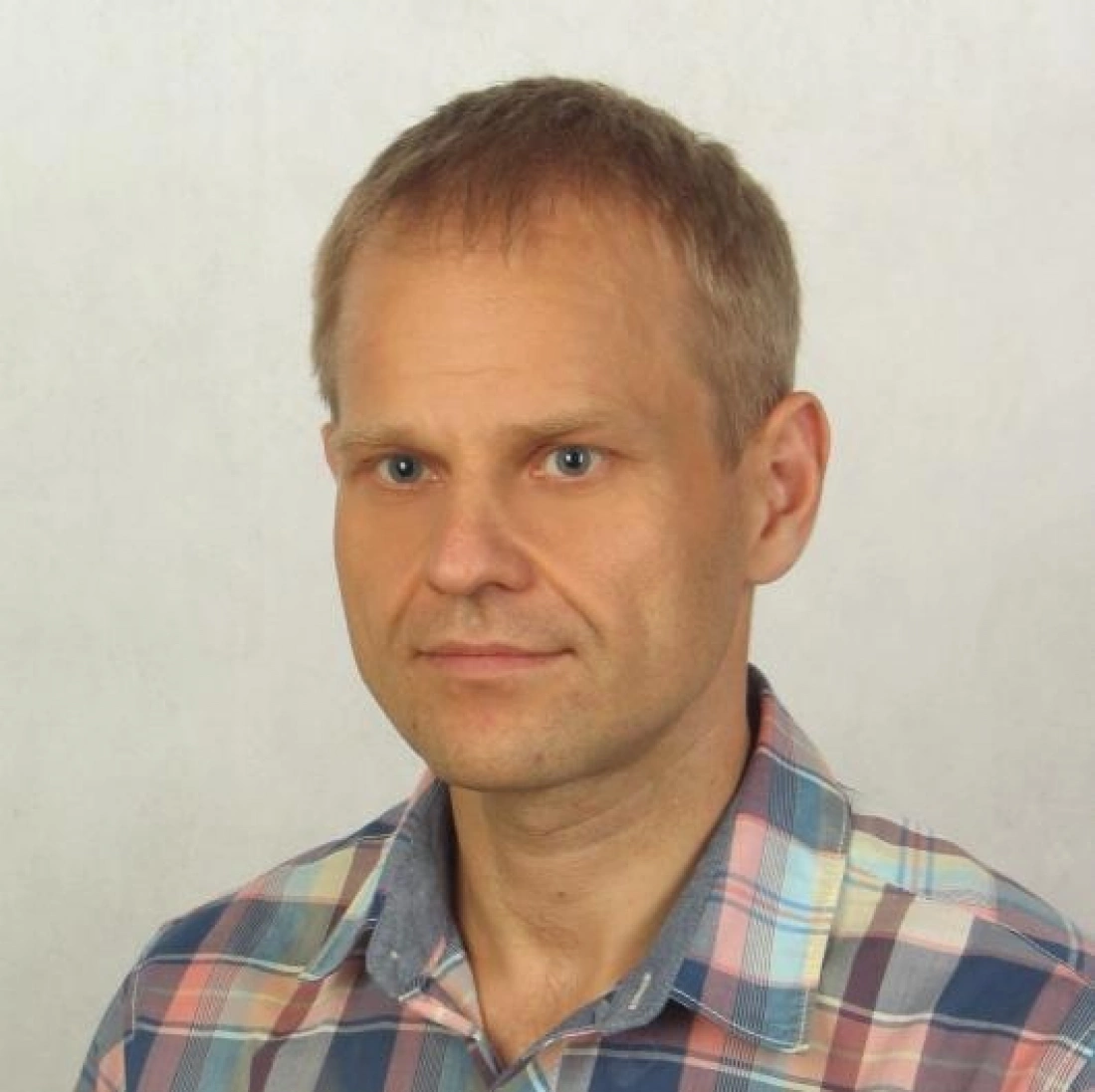 Mężczyzna w koszuli w kratkę, z neutralnym wyrazem twarzy, stojący na tle jasnej ściany.