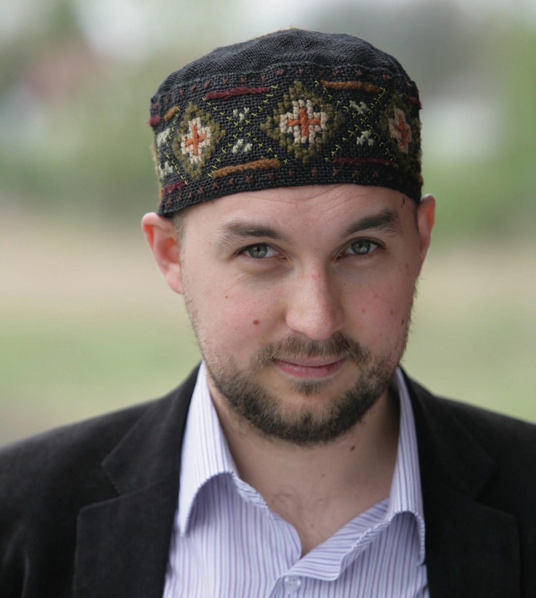 Mężczyzna w czarnej czapce z kolorowym wzorem i marynarce, patrzy w obiektyw.