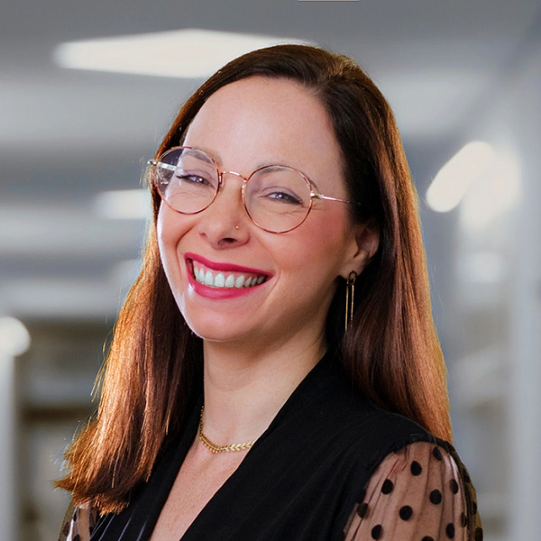 Uśmiechnięta kobieta z długimi, brązowymi włosami, nosząca okulary i elegancką bluzkę, pozująca w jasnym wnętrzu.