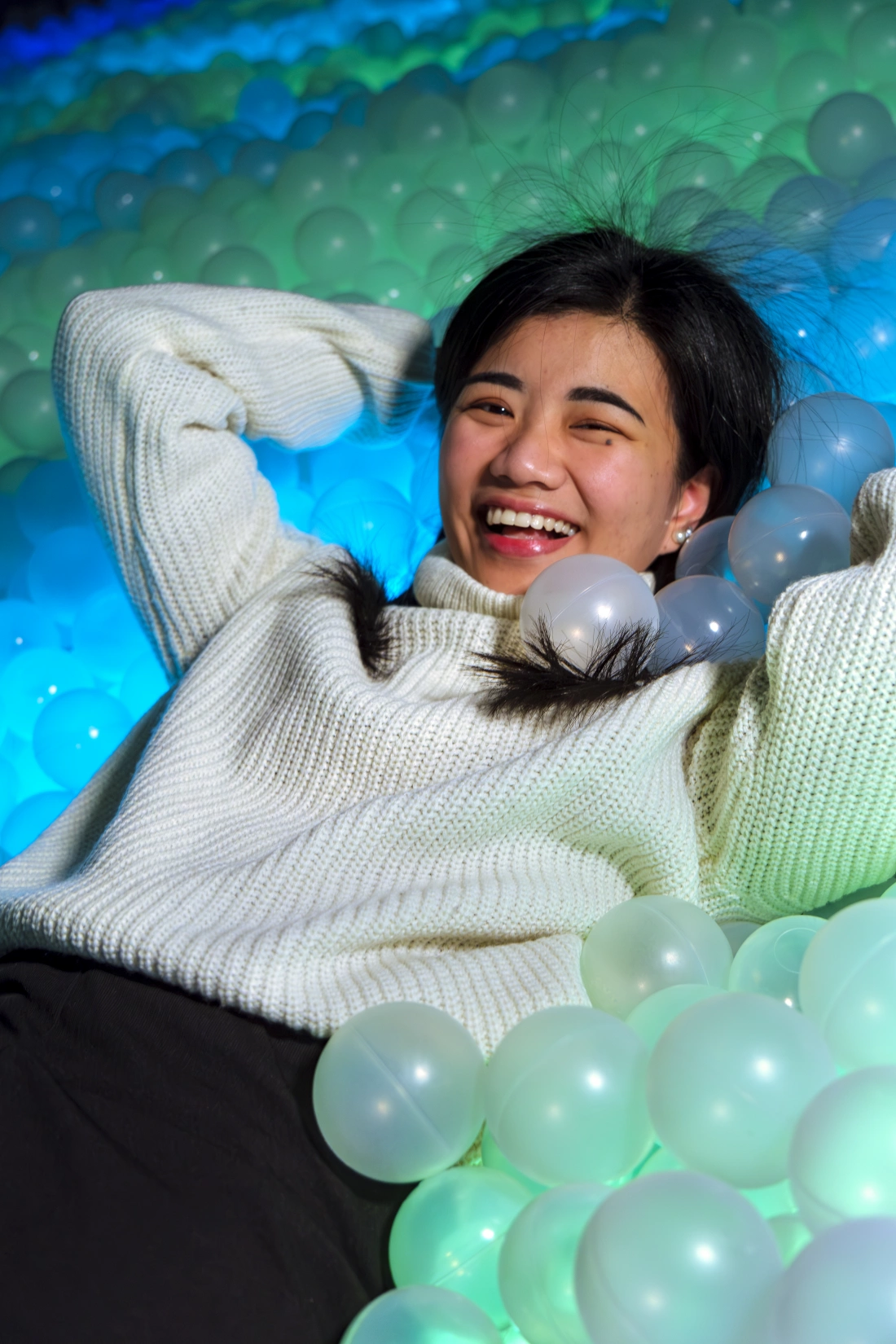 Młoda kobieta leży w kolorowych piłkach i uśmiecha się, otoczona błękitnym i zielonym tłem.