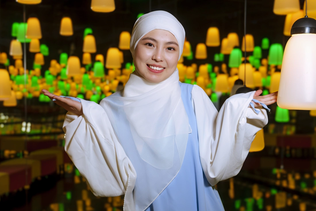 Kobieta w białym hijabie uśmiecha się i unosi ręce w geście zapraszającym, w tle kolorowe lampy.