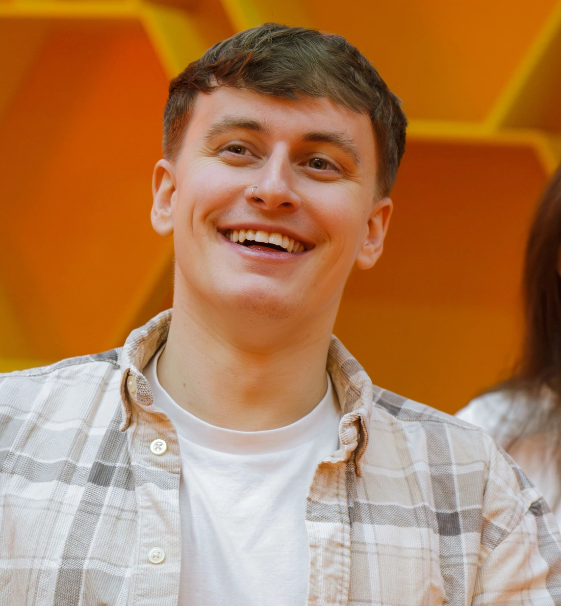 Mężczyzna z uśmiechem, ubrany w koszulę w kratkę, na tle pomarańczowej ściany.