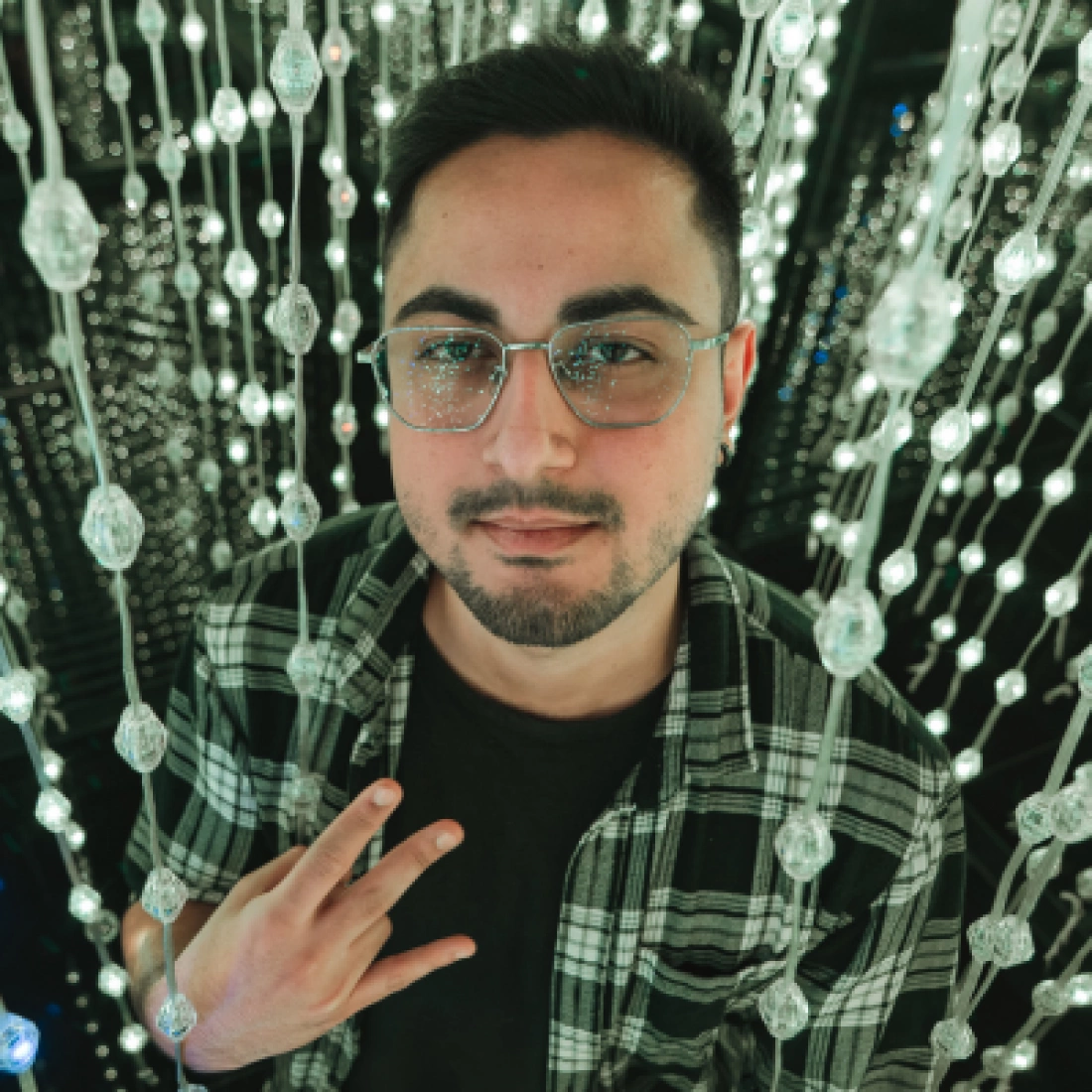 Młody mężczyzna w okularach, z plaidową koszulą, stoi pośród wiszących dekoracji świetlnych.