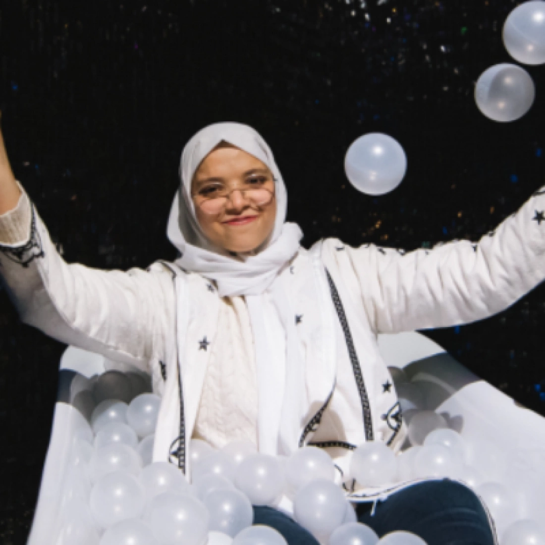 Młoda kobieta w białym szaliku i swetrze siedzi w wanny pełnej przezroczystych kul, uśmiechając się i unosi ręce w radości.