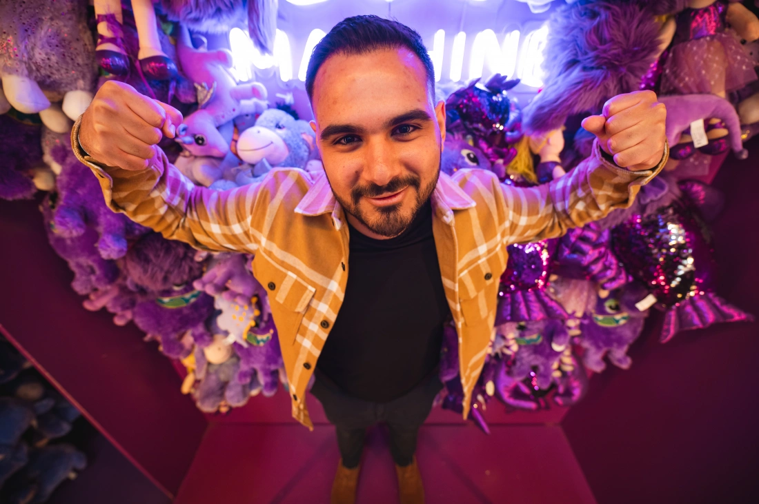 Mężczyzna w brązowej koszuli z wzorem, uśmiechający się na tle kolorowych pluszaków.