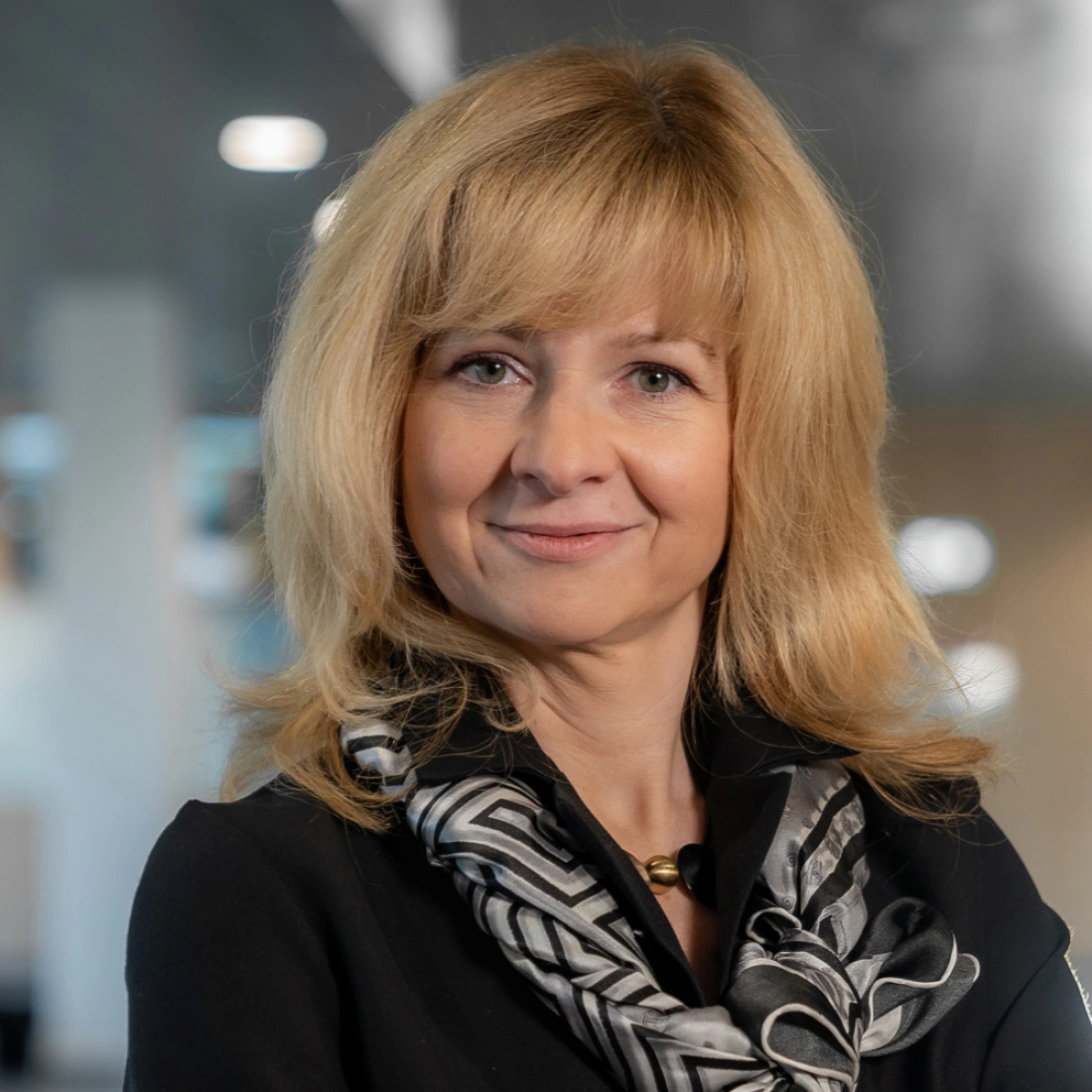 Kobieta z włosami w odcieniu blond, uśmiechająca się, w czarnym ubraniu i apaszce, na tle rozmytej przestrzeni.