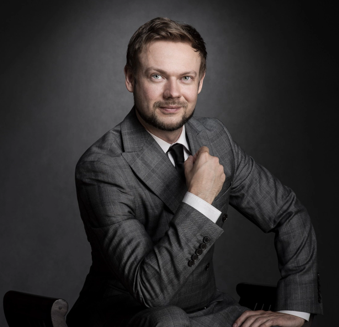 Mężczyzna w szarym garniturze, siedzący na krześle, z uśmiechem i pewną postawą.