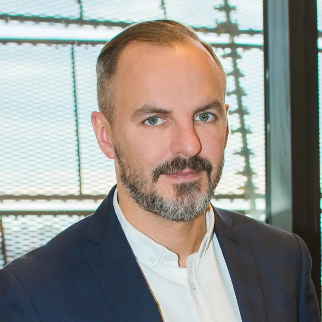 Mężczyzna z brodą w eleganckim garniturze, stojący w nowoczesnym wnętrzu.