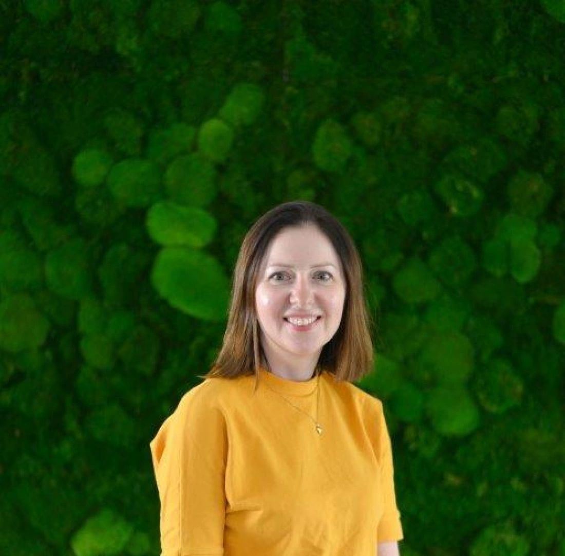 Kobieta w żółtej bluzce stoi na tle zielonej, roślinnej kompozycji.