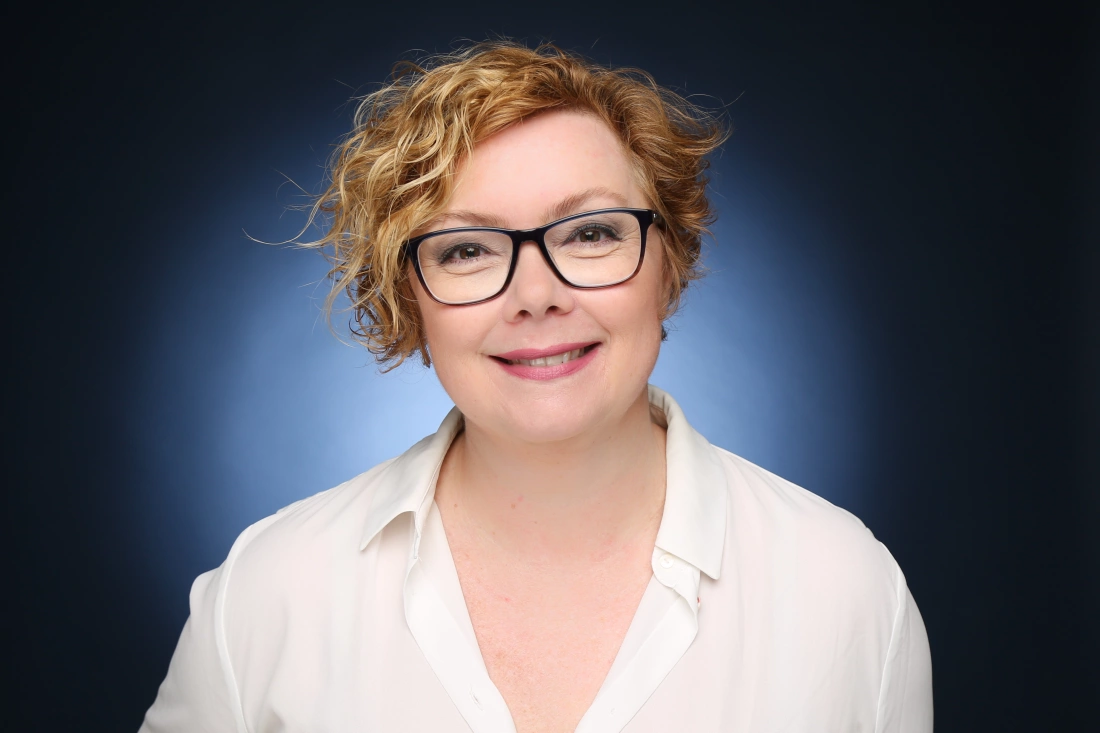 Kobieta z krótkimi, kręconymi włosami, w okularach, uśmiechająca się na zdjęciu. Ubrana w białą koszulę, na tle w odcieniach niebieskiego.