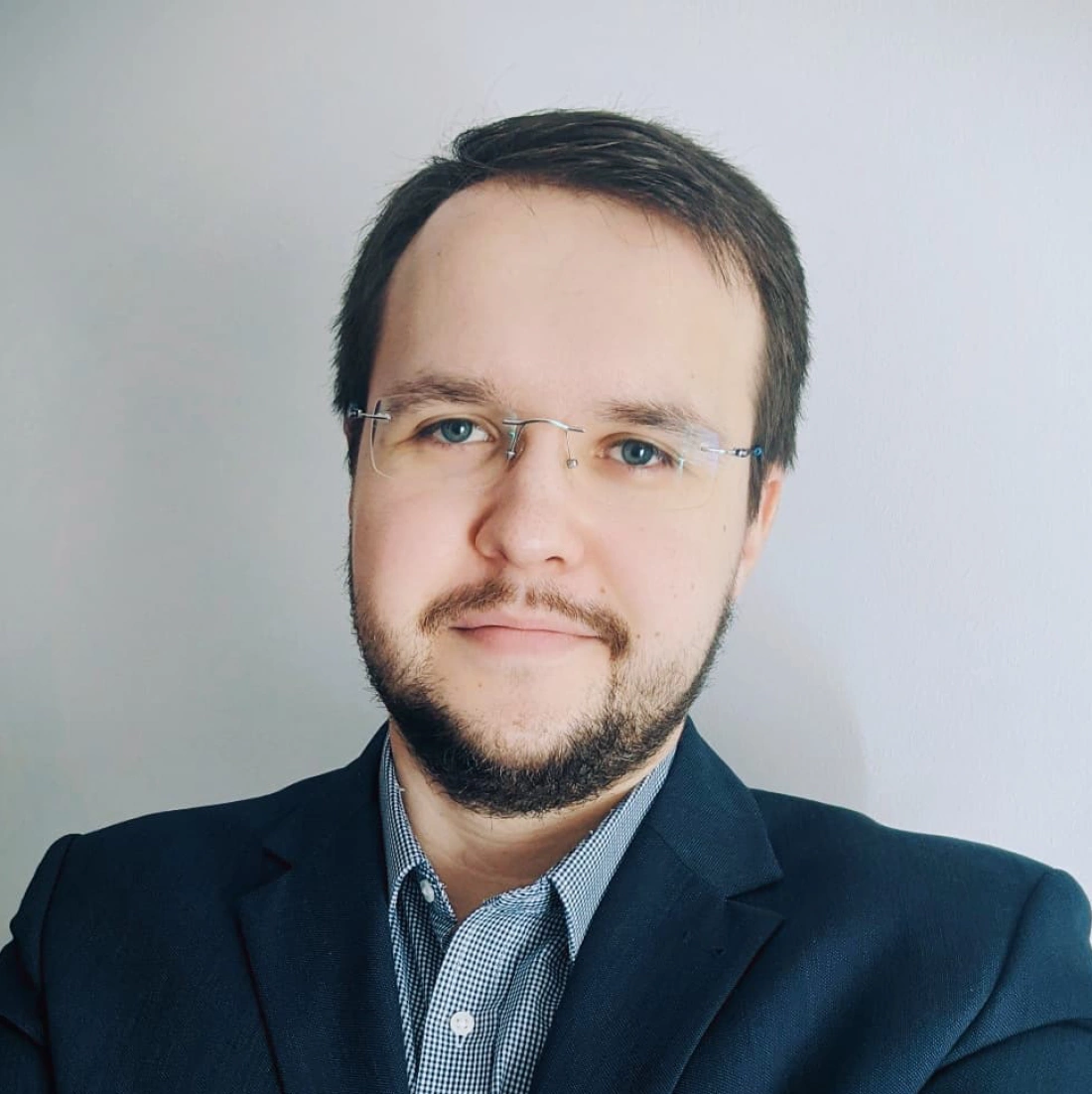 Mężczyzna w okularach, uśmiechający się na tle jasnej ściany, ubrany w garnitur.