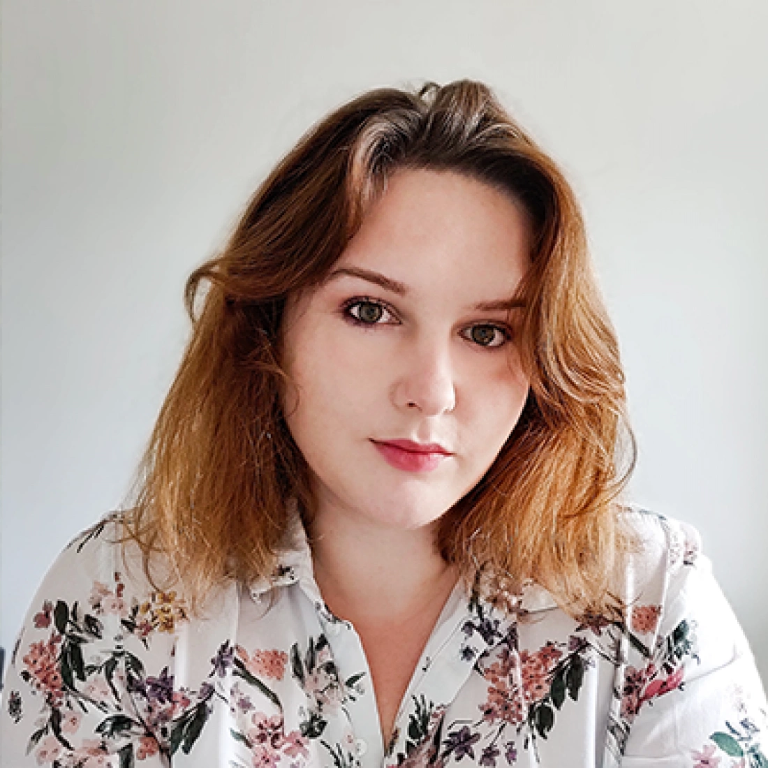 Młoda kobieta z brązowymi włosami, ubrana w koszulę w kwiaty, patrzy w kamerę.
