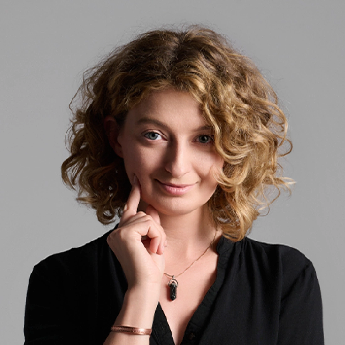 Młoda kobieta z kręconymi włosami, uśmiechająca się i wskazująca palcem na policzek.