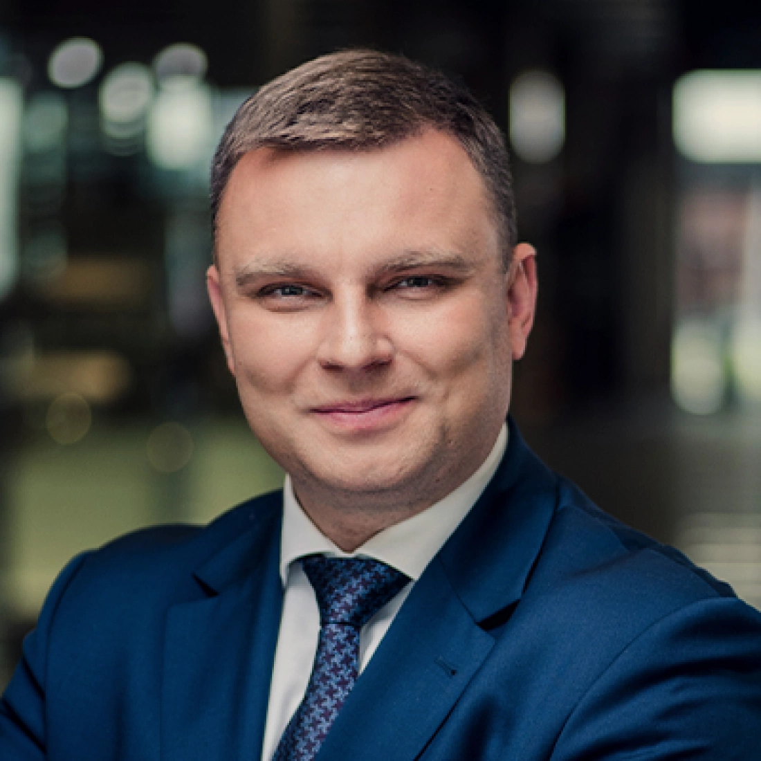 Mężczyzna w garniturze, uśmiechający się do kamery, w tle zamazane biuro.