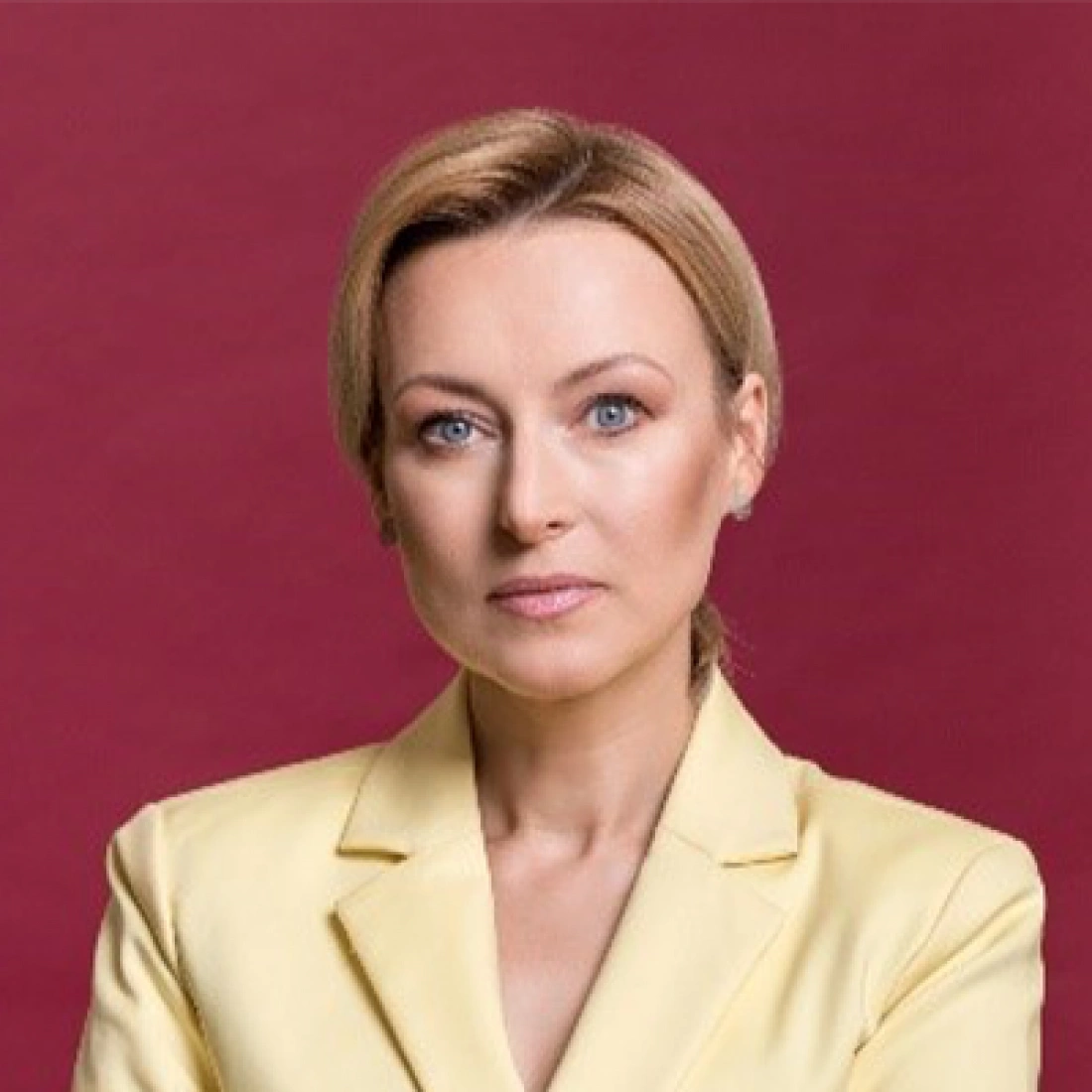 Kobieta w żółtej marynarce, z poważnym wyrazem twarzy, na tle w odcieniu burgunda.