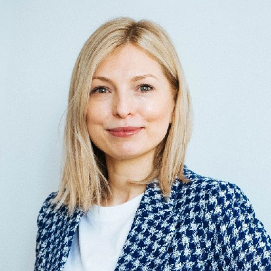 Młoda kobieta z blond włosami, ubrana w niebieską marynarkę w kratę, uśmiecha się.