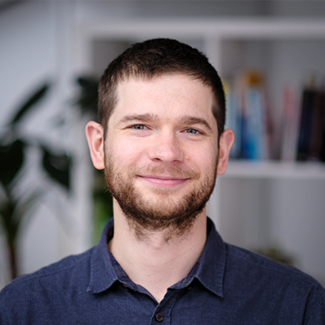 Mężczyzna z brodą, uśmiechający się, stoi przed półką z książkami.