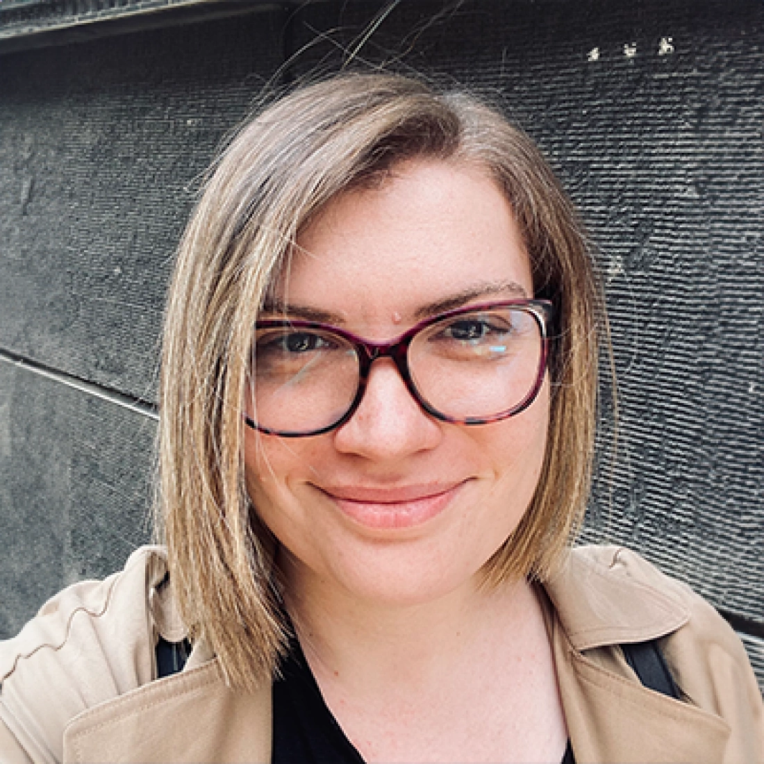 Młoda kobieta z okularami i krótkimi, prostymi włosami, uśmiecha się na tle szarej ściany.