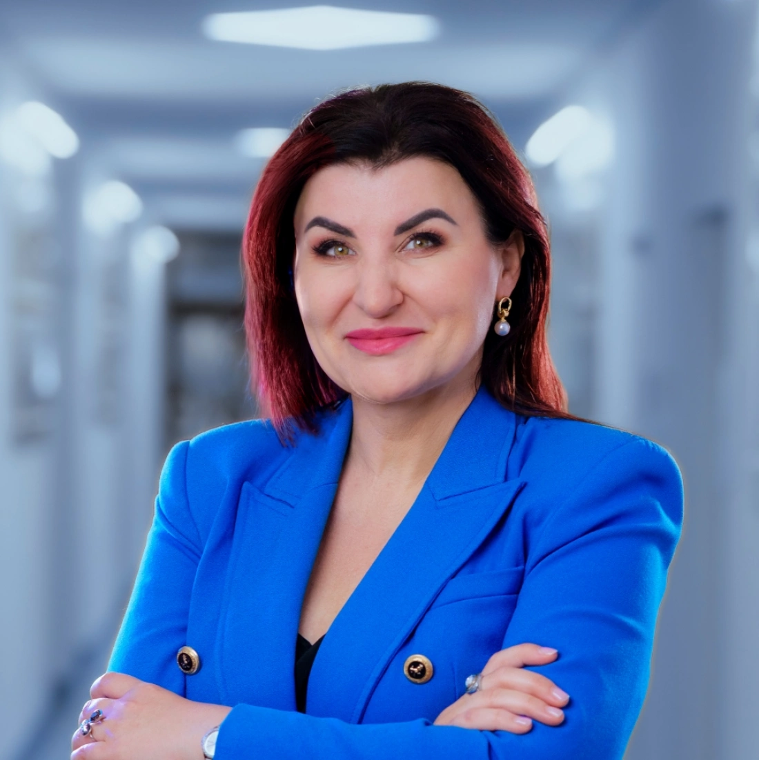 Kobieta w niebieskiej marynarce z uśmiechem, stojąca z założonymi rękami na tle jasnego korytarza.