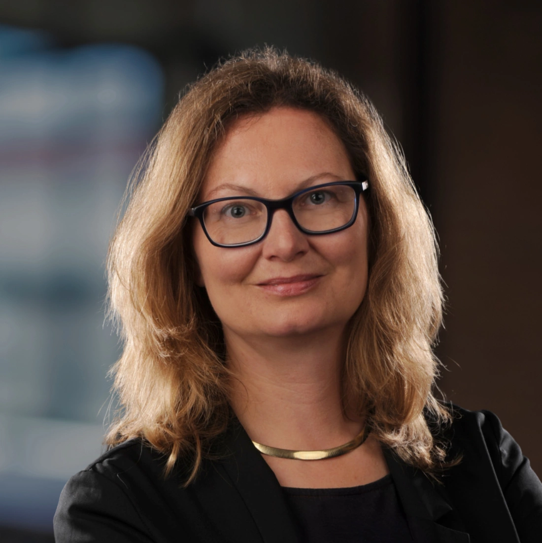 Kobieta z długimi, falującymi włosami, nosząca okulary i czarną marynarkę, uśmiecha się w profesjonalnej scenerii.