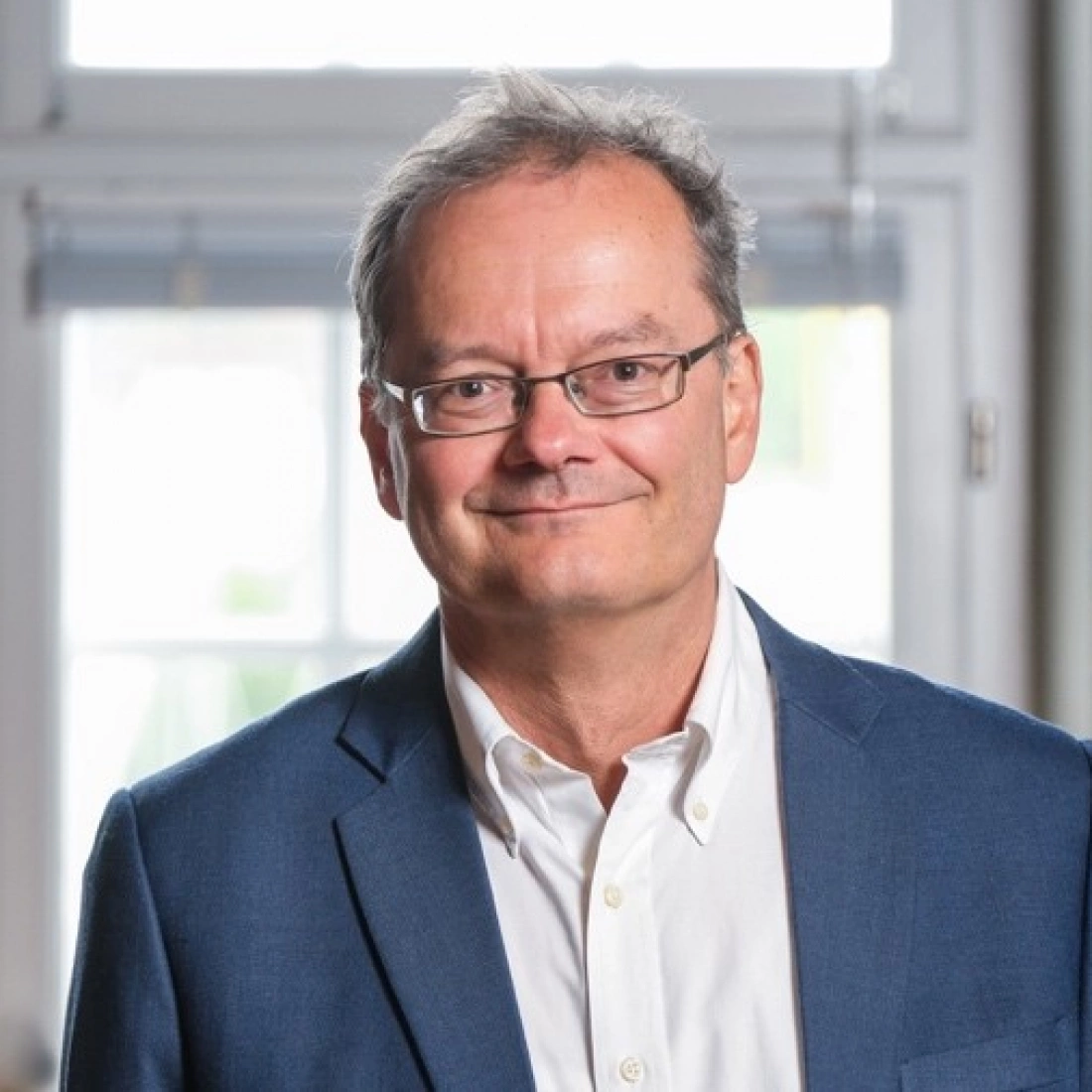 Mężczyzna w okularach w garniturze, uśmiechający się przed jasnym tłem.