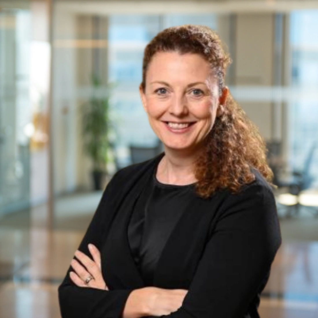 Uśmiechnięta kobieta w czarnym ubraniu, z rękami skrzyżowanymi na piersi, stoi w biurze.