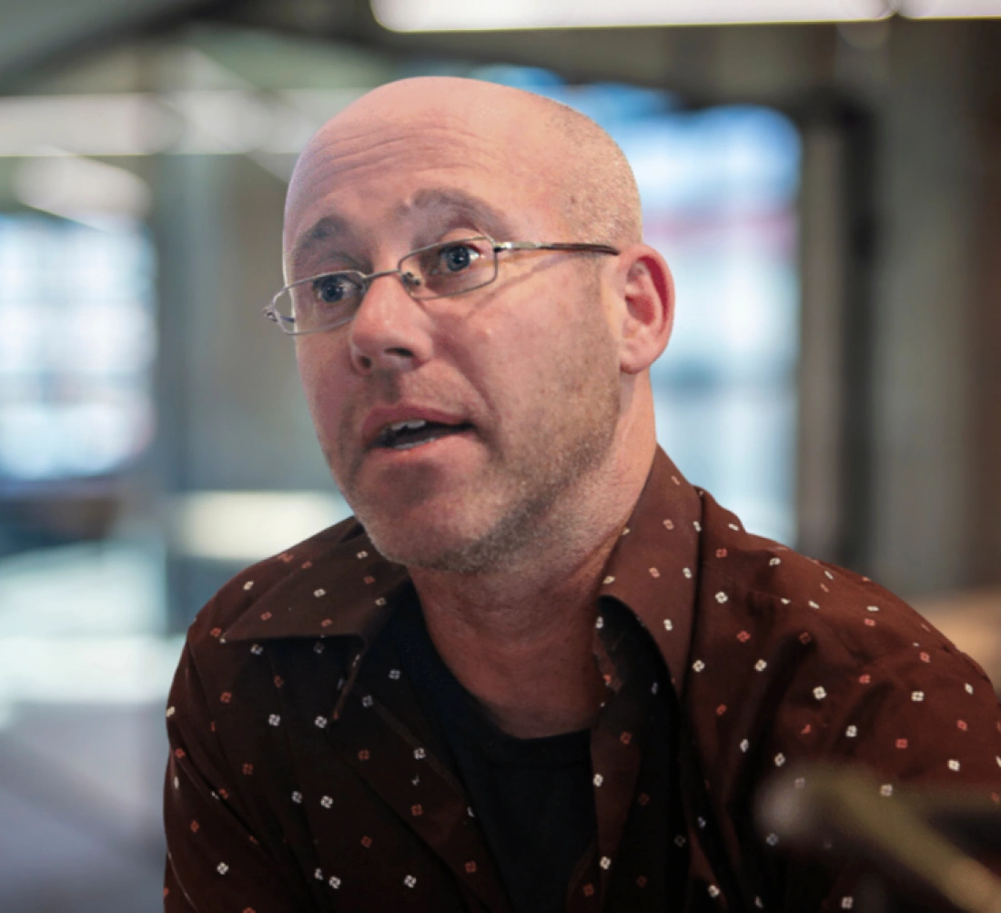 Mężczyzna z okularami, krótko ostrzyżony, w brązowej koszuli z wzorem, rozmawia w tle nowoczesnego wnętrza.