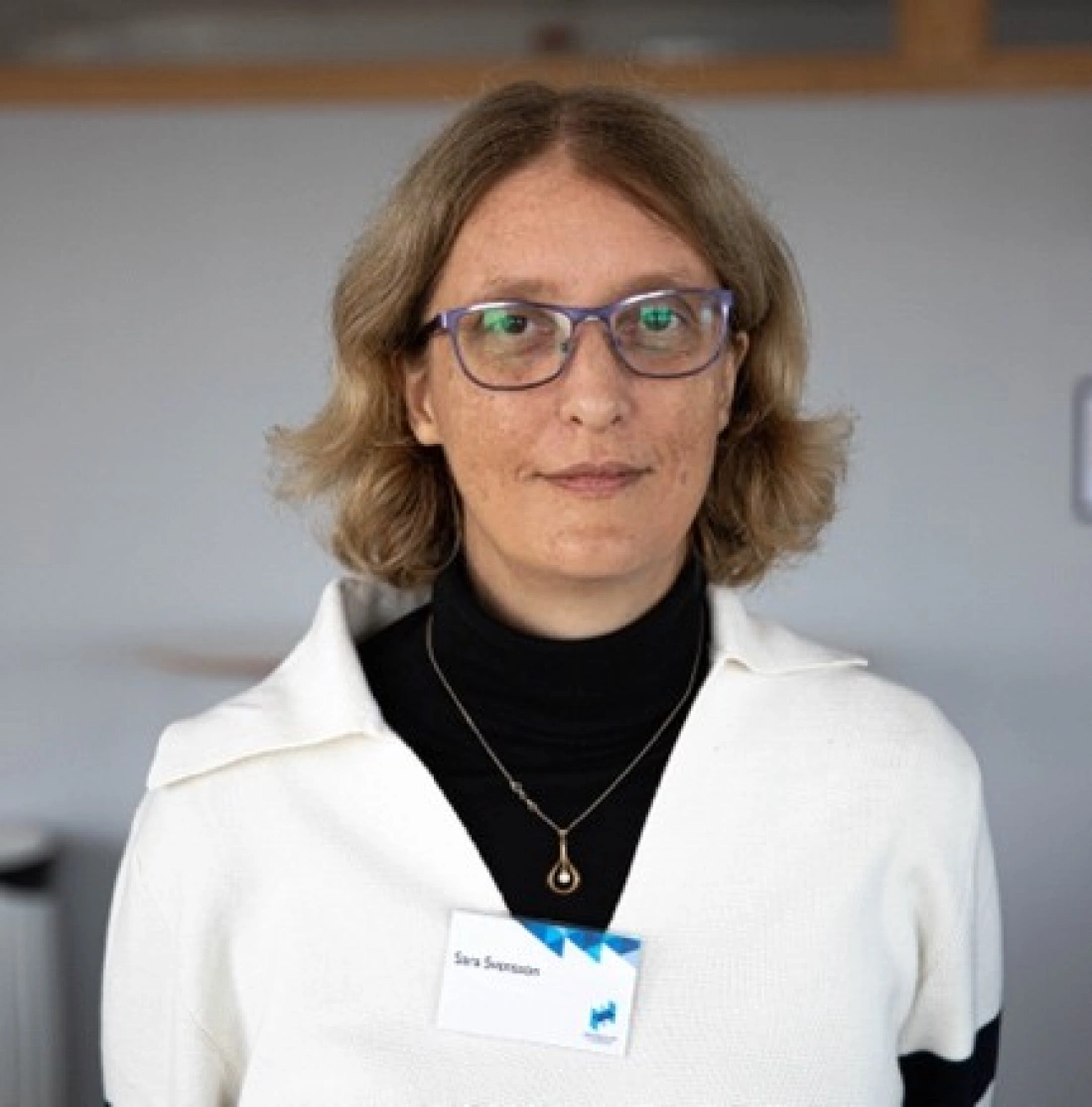 Kobieta z krótkimi, falowanymi włosami, nosząca okulary, ubrana w biały sweter i czarną bluzkę. W tle widać jasne otoczenie biurowe.