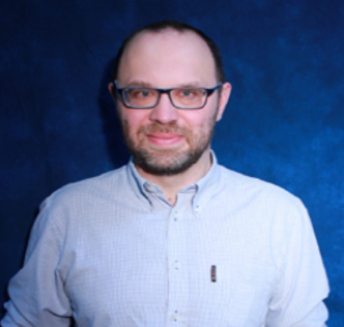 Mężczyzna w okularach, ubrany w jasną koszulę, stoi przed niebieskim tłem.