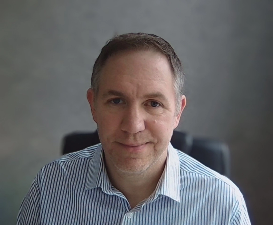 Mężczyzna w niebieskiej koszuli w paski siedzi przed szarym tłem.