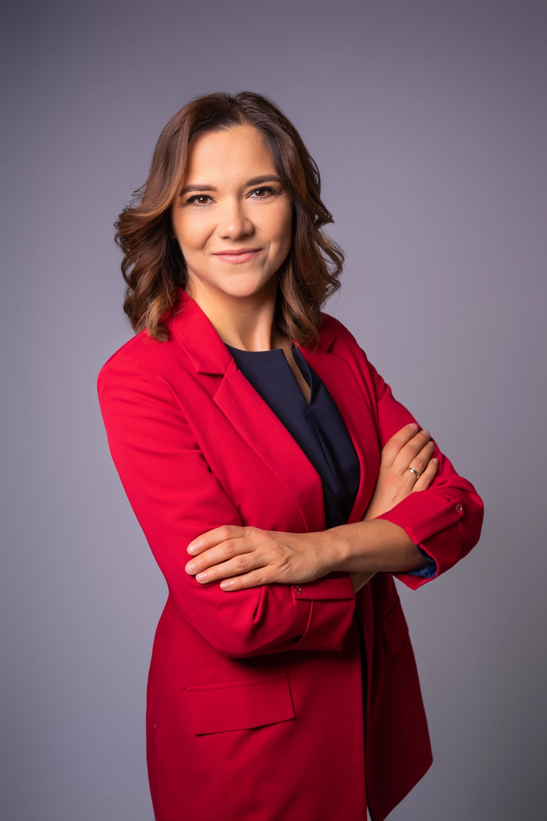 Kobieta w czerwonym żakiecie z założonymi rękami, uśmiechająca się. Tło w odcieniach szarości.