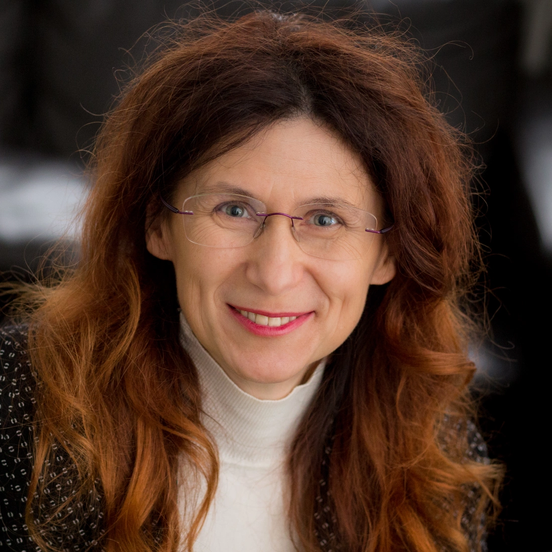 Uśmiechnięta kobieta z długimi, falującymi włosami, w okularach, w białej bluzce.