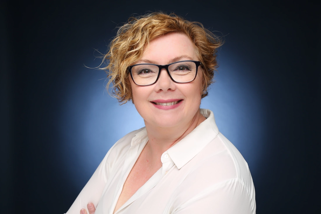 Portret uśmiechającej się kobiety w okularach, w białej bluzce, na ciemnym tle.