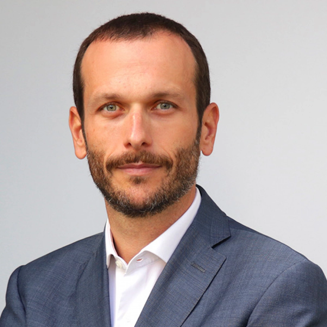 Mężczyzna w garniturze z krótkimi włosami i zarostem, stojący na neutralnym tle.