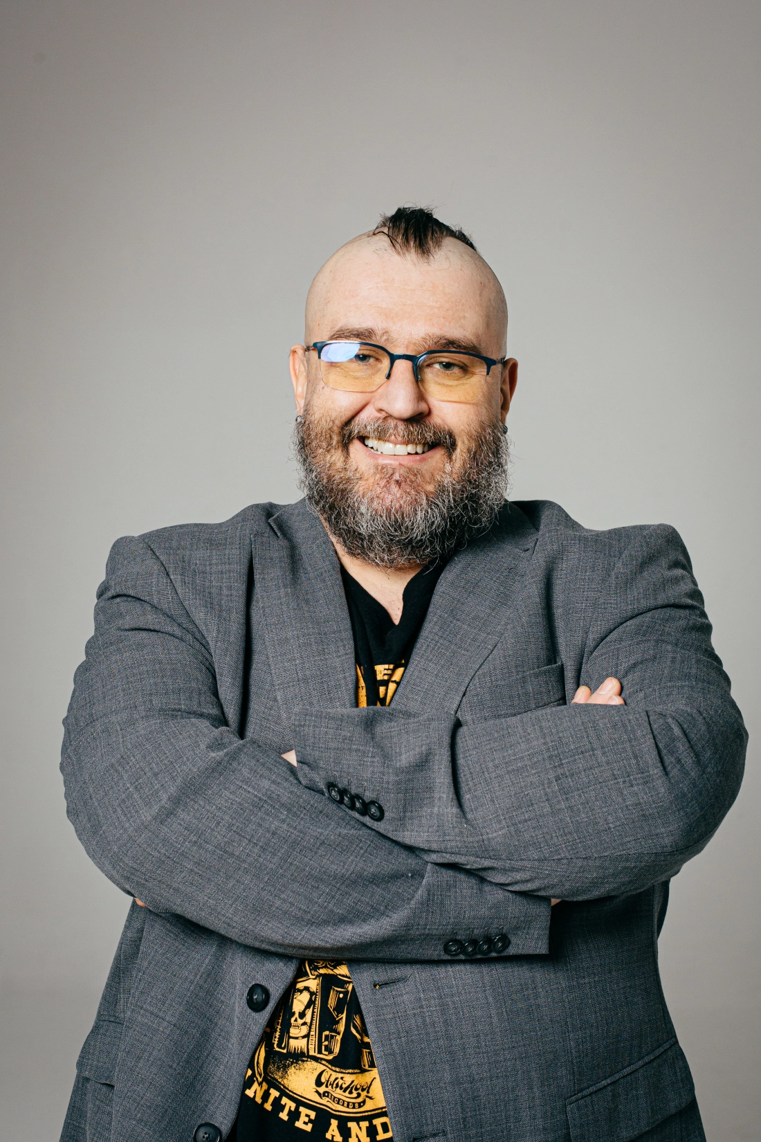 Mężczyzna z brodą, w okularach, uśmiechający się w szarym garniturze, z rękami skrzyżowanymi na piersi.