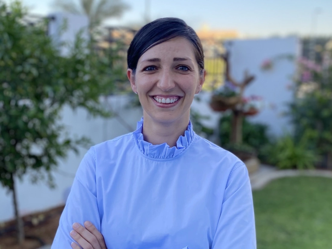 Uśmiechnięta kobieta w niebieskiej bluzie, stojąca na tle zieleni.