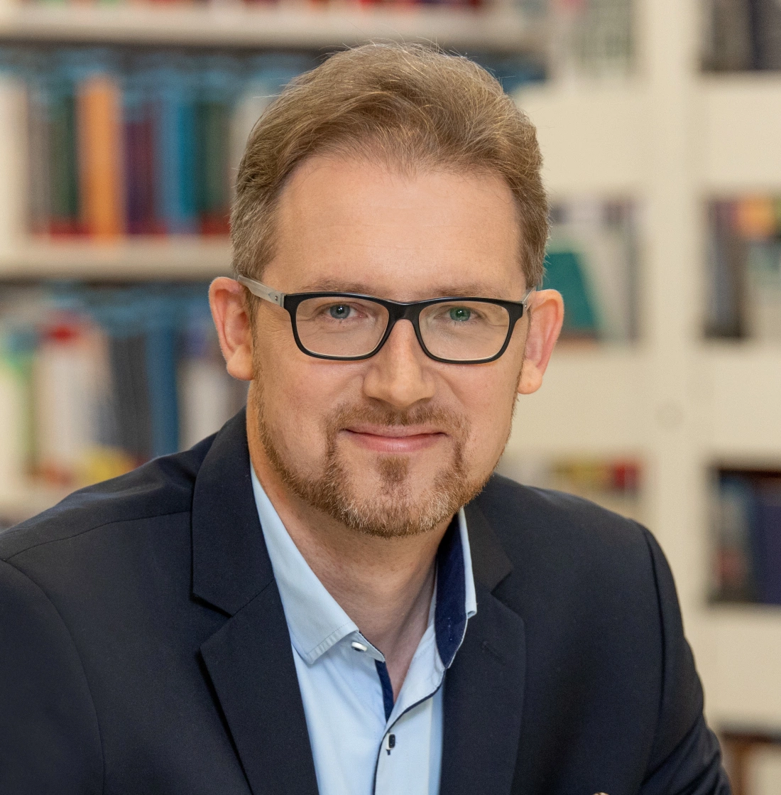 Mężczyzna w okularach uśmiecha się, siedząc w bibliotece, w tle widoczne półki z książkami.