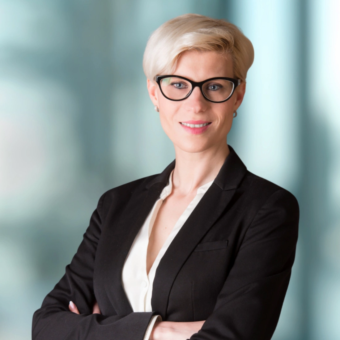 Kobieta w okularach, ubrana w czarną marynarkę, z założonymi rękami, uśmiechająca się. Tło w odcieniach niebieskiego.