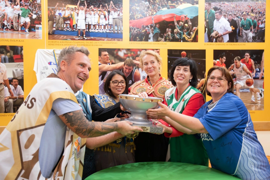 Grupa osób uśmiecha się, trzymając puchar, w tle zdjęcia związane z koszykówką.