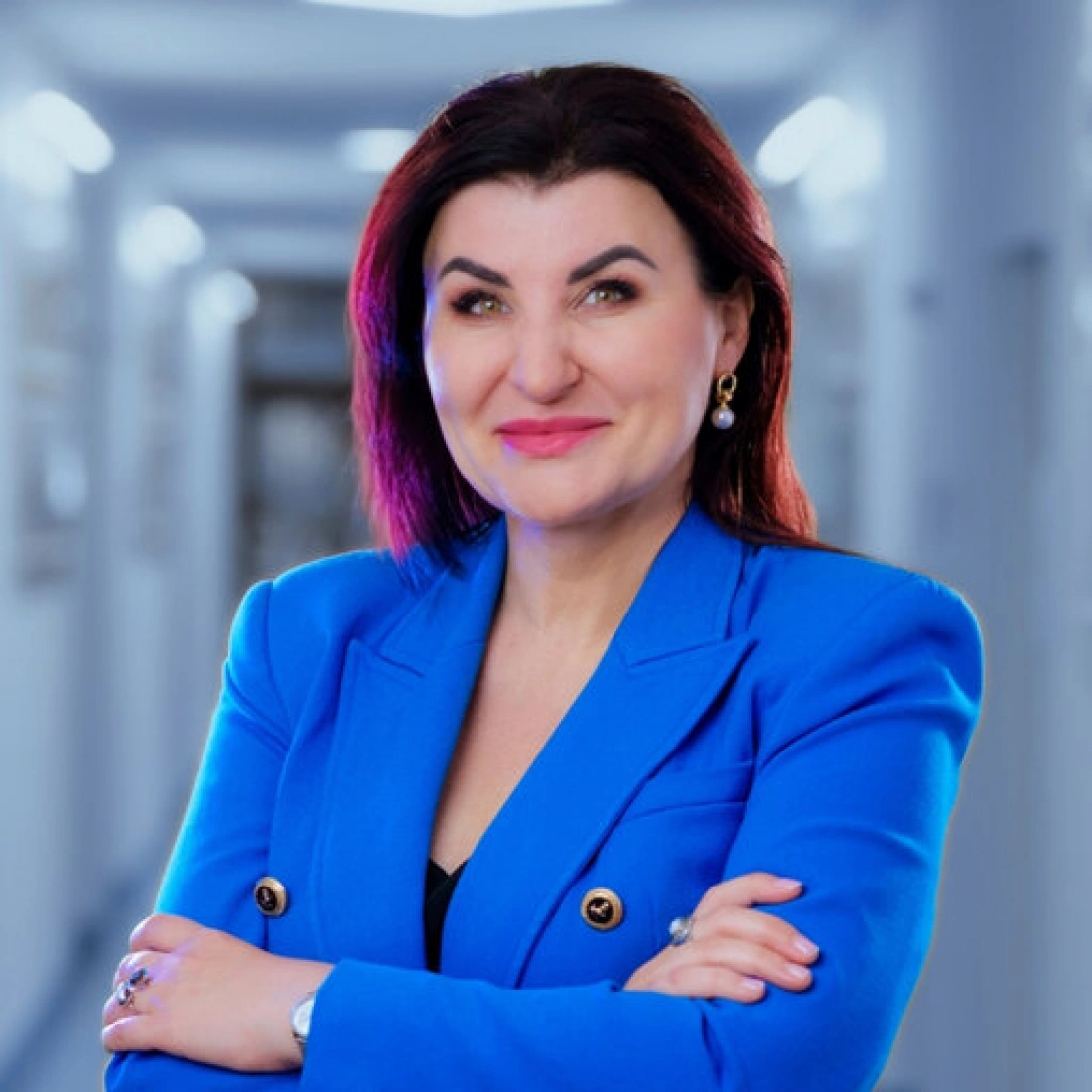 Kobieta w niebieskiej marynarce, uśmiecha się i ma założone ręce, w tle jasny korytarz.