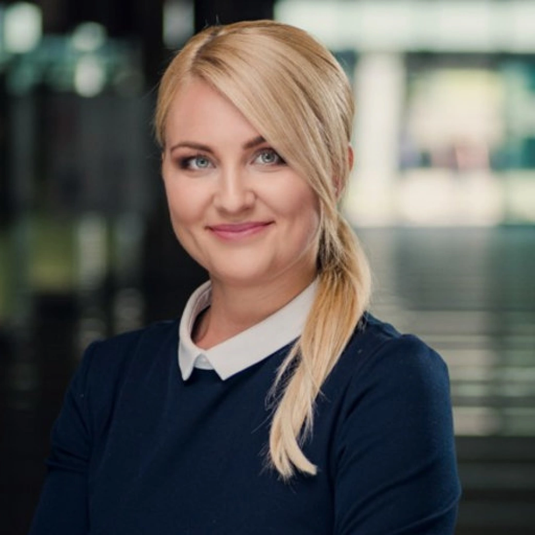 Kobieta z blond włosami, uśmiechająca się, w eleganckim stroju, stojąca na tle rozmytej przestrzeni.