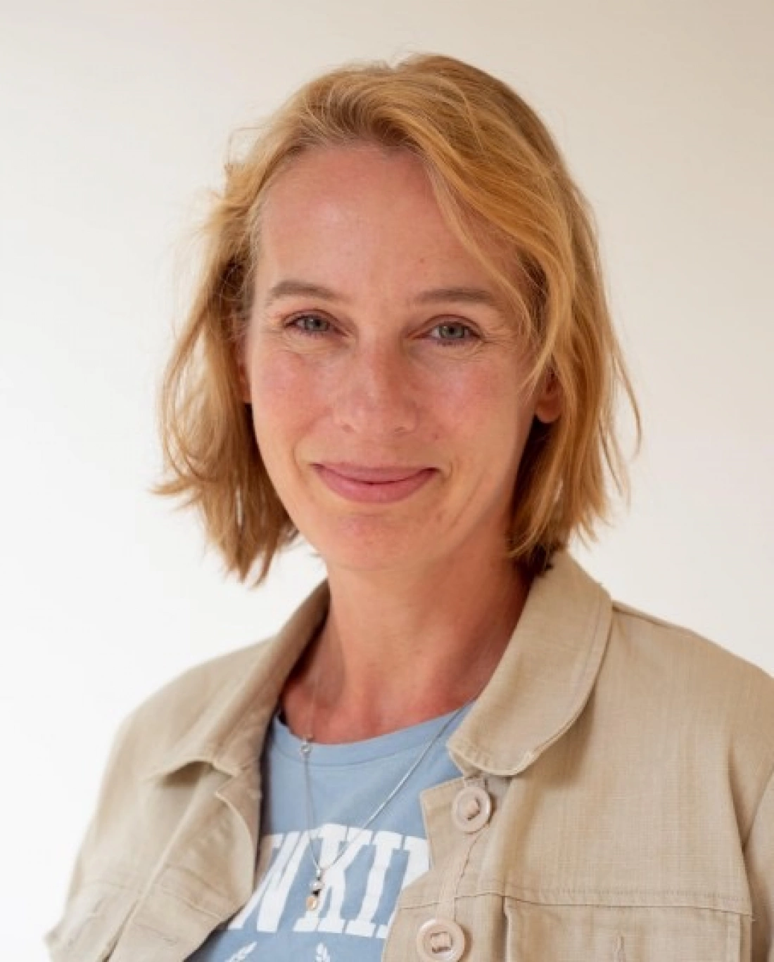 Kobieta z krótkimi, blond włosami, uśmiechająca się na neutralnym tle.