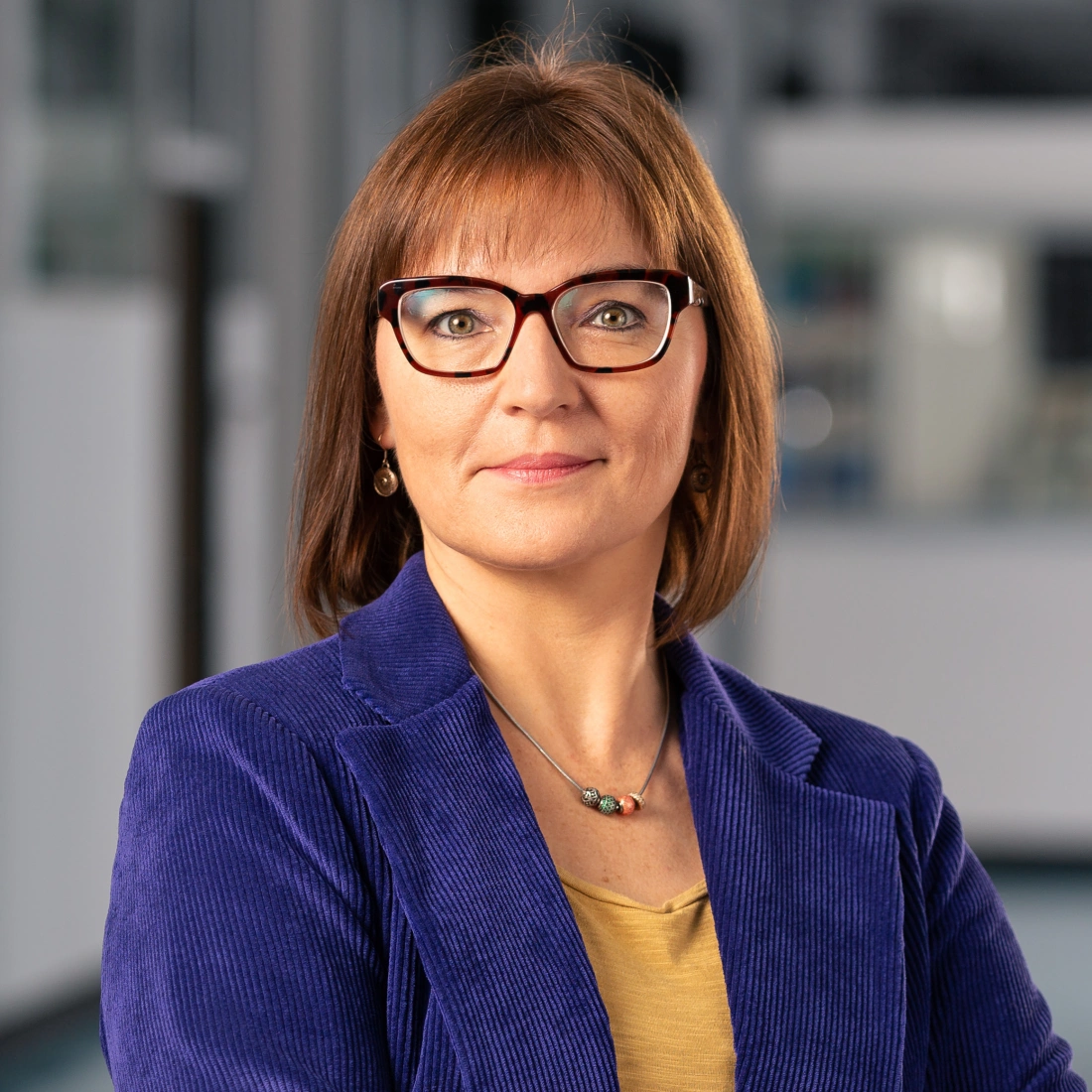 Zdjęcie przedstawia kobietę w okularach, ubraną w granatową marynarkę, z lekkim uśmiechem na tle nowoczesnego wnętrza.