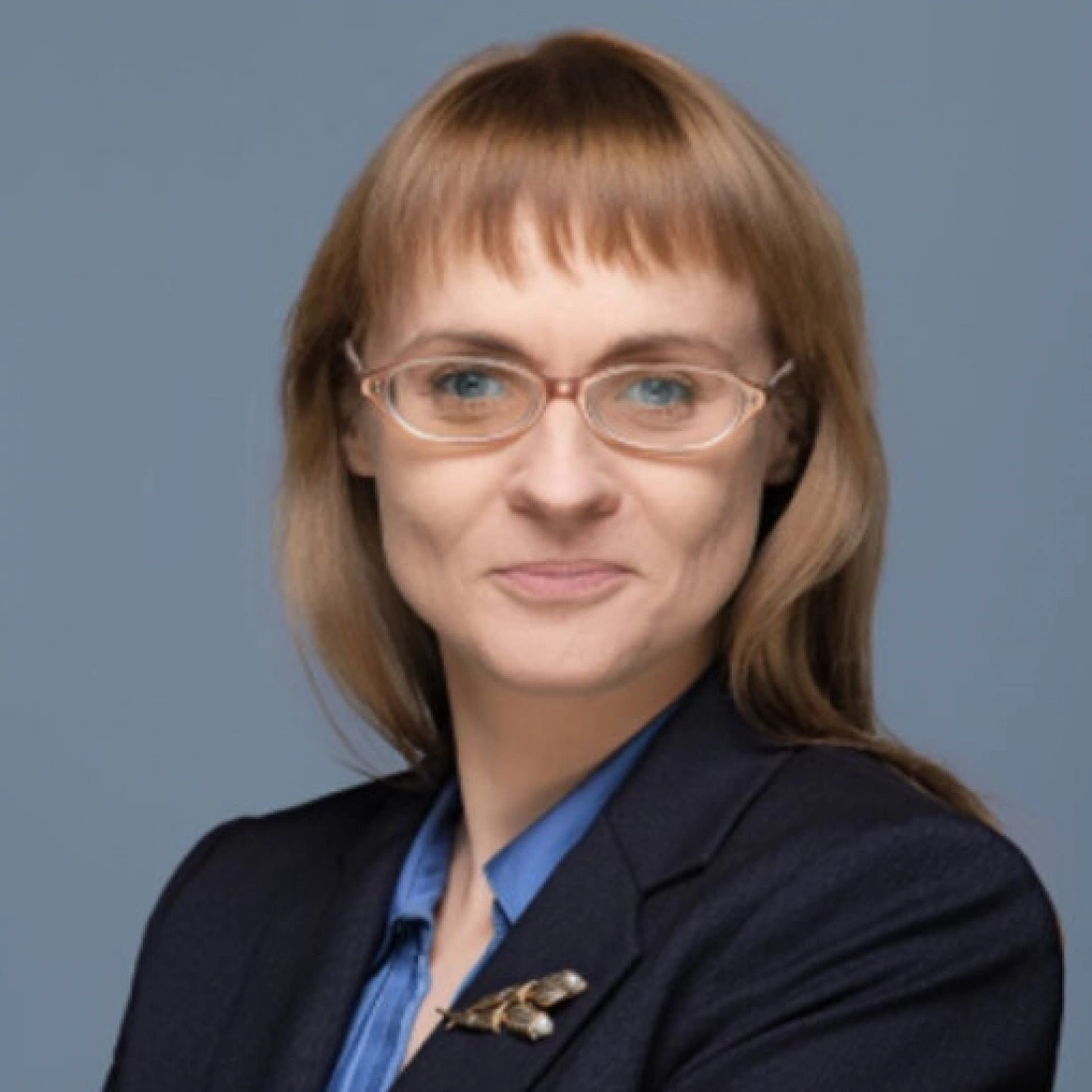 Kobieta o krótkich włosach, w okularach, w eleganckiej marynarce, uśmiecha się do kamery.