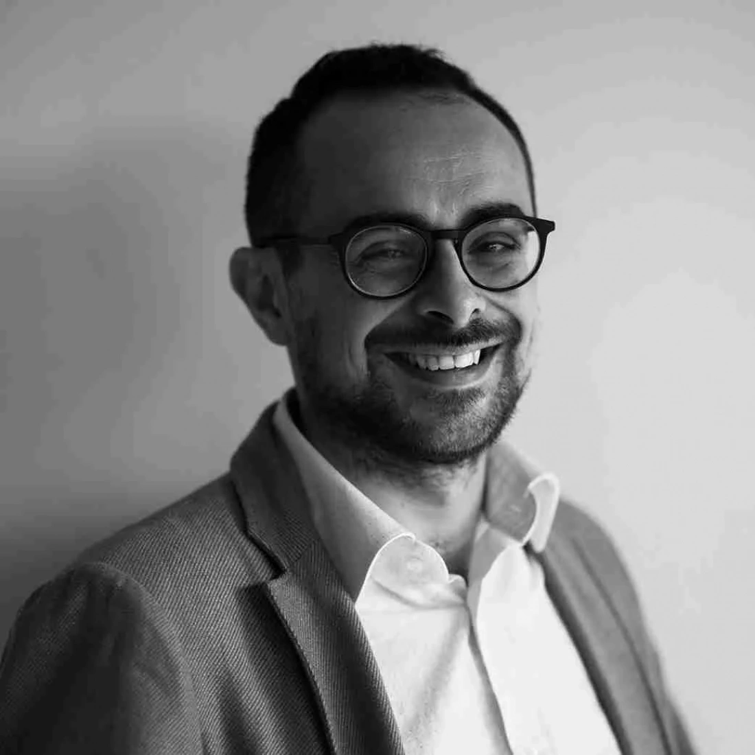 Uśmiechnięty mężczyzna w okularach, ubrany w garnitur, na neutralnym tle.