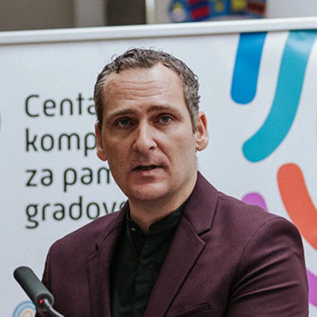Mężczyzna w eleganckim ubraniu mówi do mikrofonu, w tle widoczny plakat z kolorowymi wzorami.