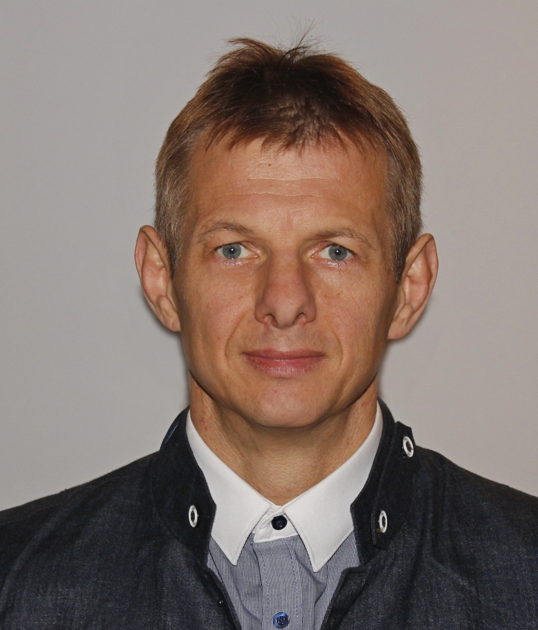 Mężczyzna z krótkimi, jasnymi włosami i niebieskimi oczami, ubrany w ciemną kurtkę i białą koszulę.