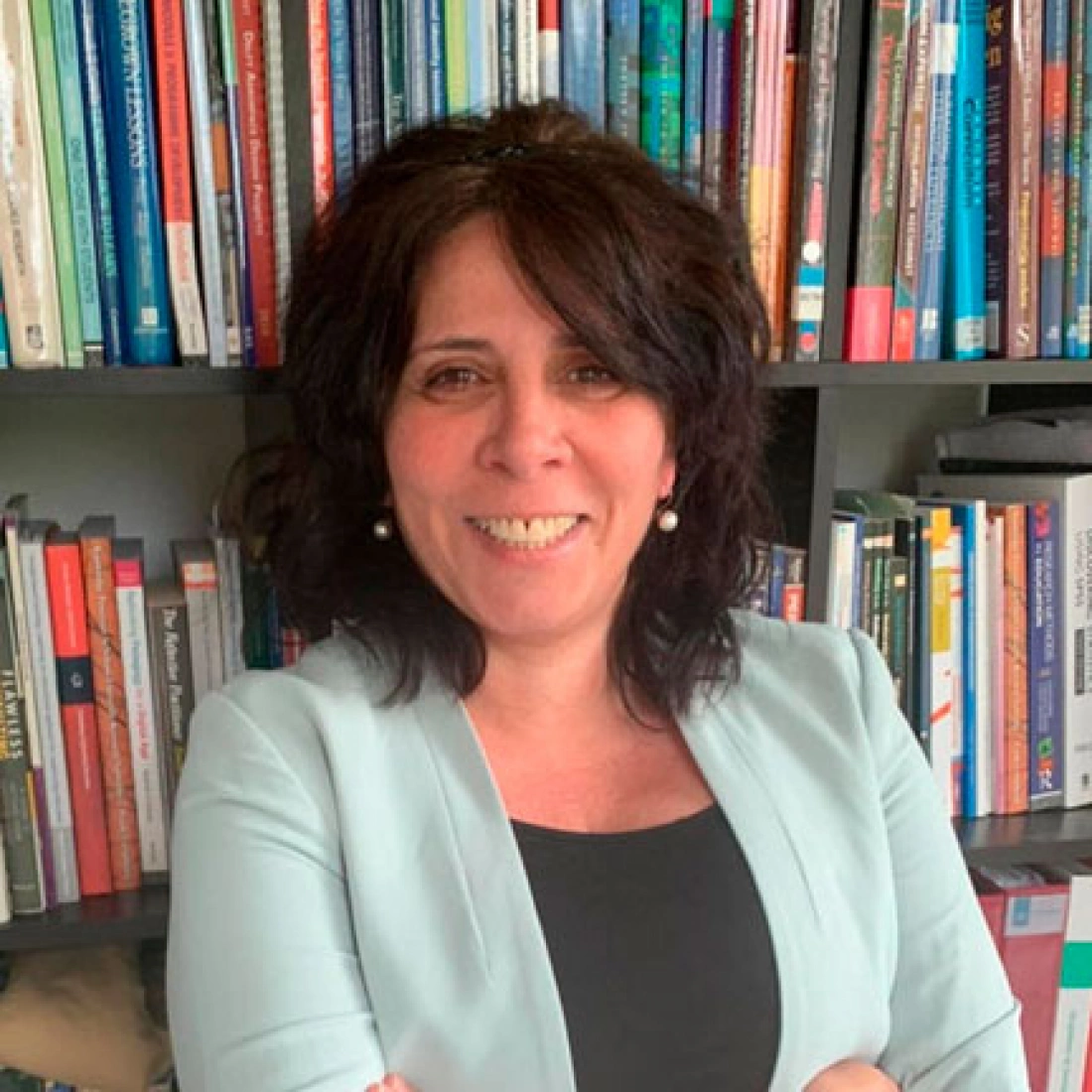 Kobieta w jasnym żakiecie stoi z uśmiechem na tle półki z książkami.