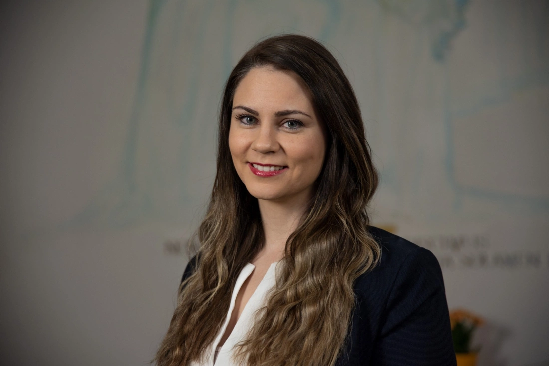 Portret młodej kobiety z długimi, falującymi włosami, uśmiechającej się w eleganckim ubraniu. W tle delikatne elementy dekoracyjne.