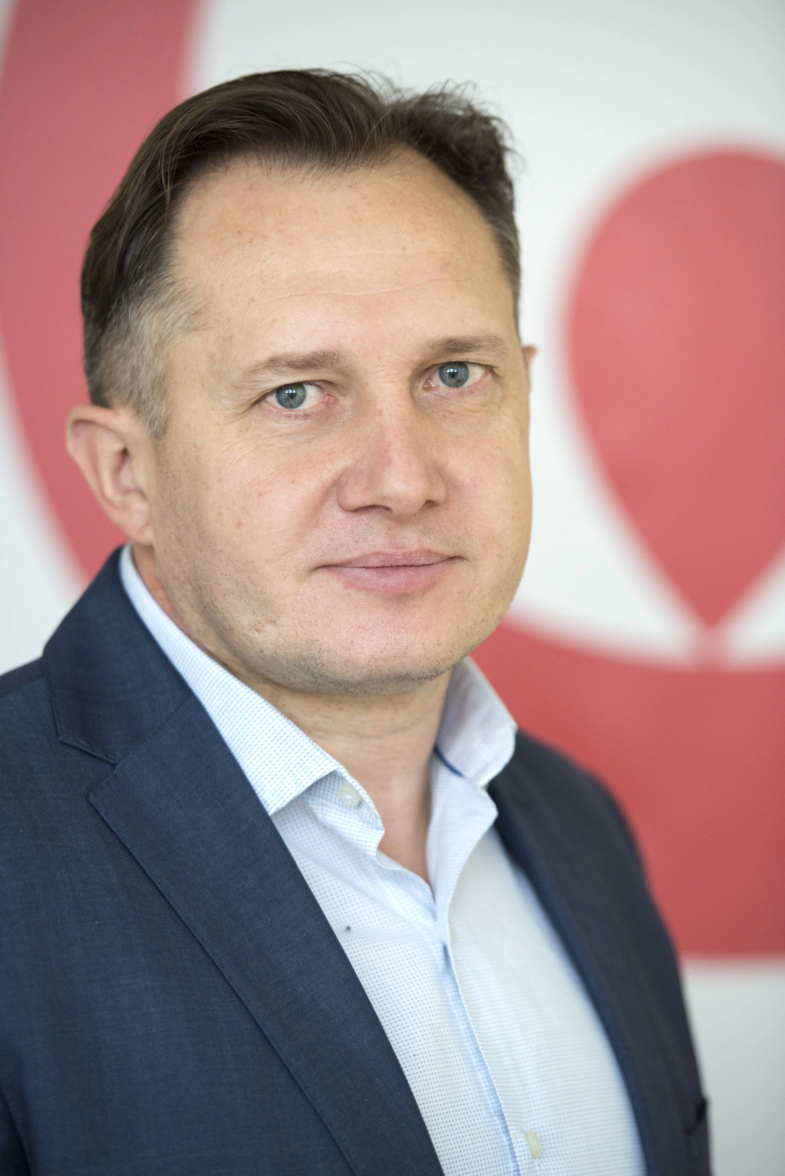 Mężczyzna w garniturze, z krótkimi włosami i niebieskimi oczami, prezentuje się na tle jasnego tła z czerwonym motywem.